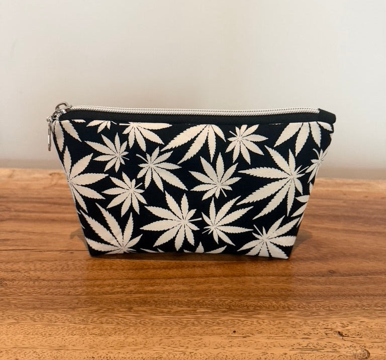 A small zippered pouch with an all-over white cannabis leaf pattern on a black background, resting on a rustic wooden surface against a plain light wall.