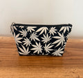 A small zippered pouch with an all-over white cannabis leaf pattern on a black background, resting on a rustic wooden surface against a plain light wall.